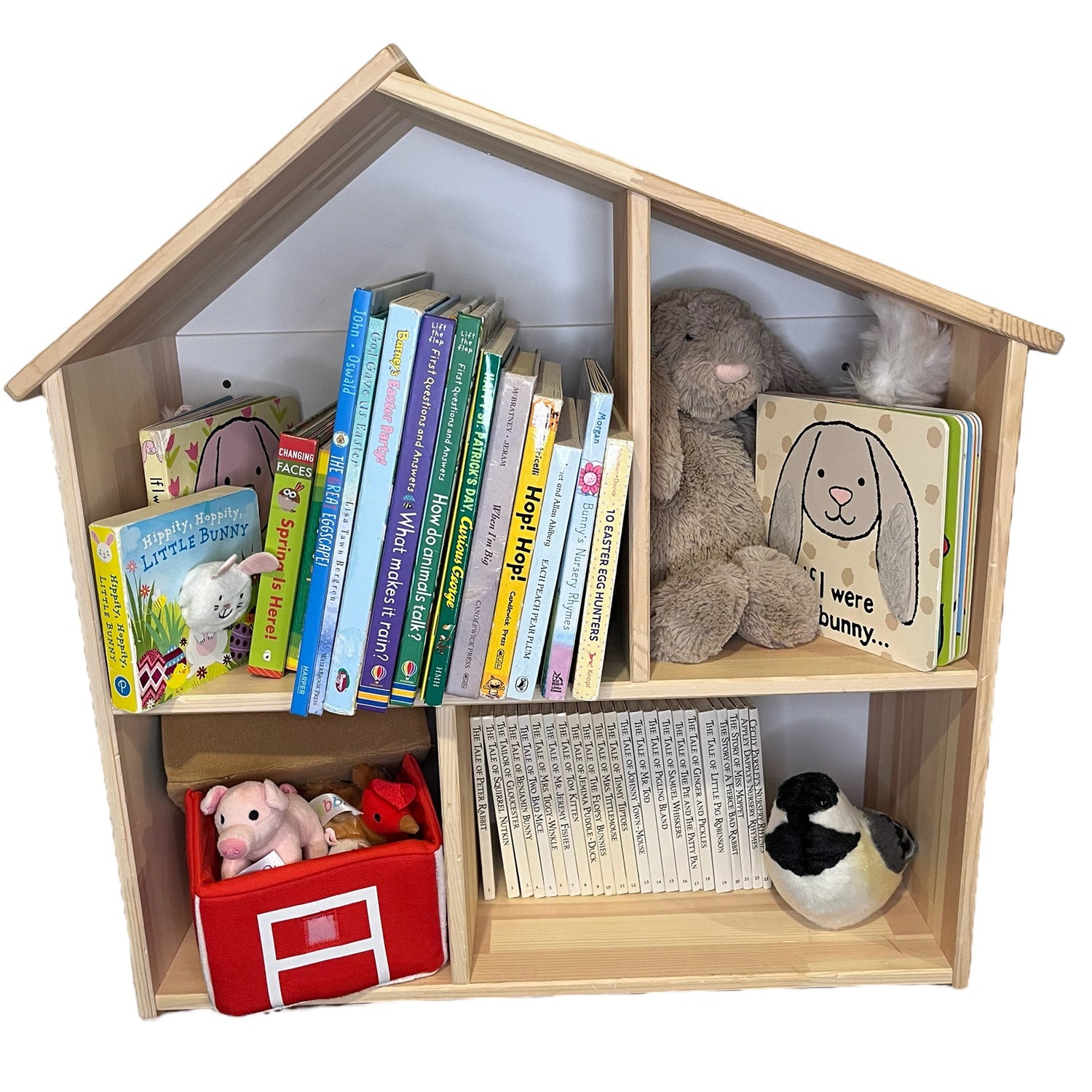 A wooden house-shaped children's bookshelf with three tiers of open shelves, styled with colorful children's books, a plush bunny, a red toy box, and a stuffed bird, set against a plain white background.